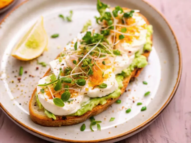 A slice of toast topped with smashed avocado, creamy cottage cheese, microgreens, and a sprinkle of spices, served with a lemon wedge on a ceramic plate.