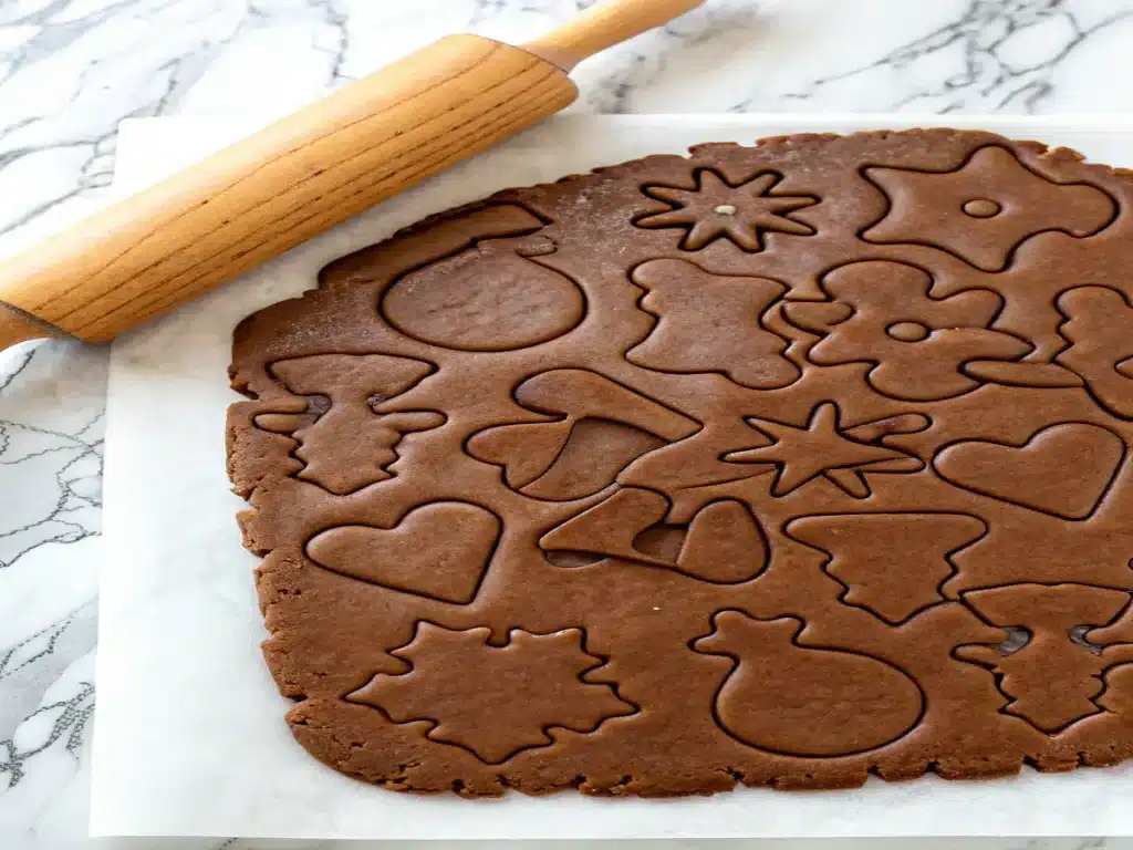 Rolled-out gingerbread dough on parchment paper, with various holiday shapes cut out, and a wooden rolling pin in the background.
