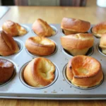 Lightly golden popovers still in their muffin tin, showing an airy texture.