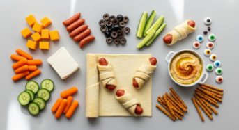 Flat lay of puff pastry, mini hot dogs, cheese, cream cheese, olives, carrots, cucumbers, hummus, pretzel sticks, and candy eyeballs on a modern kitchen counter.