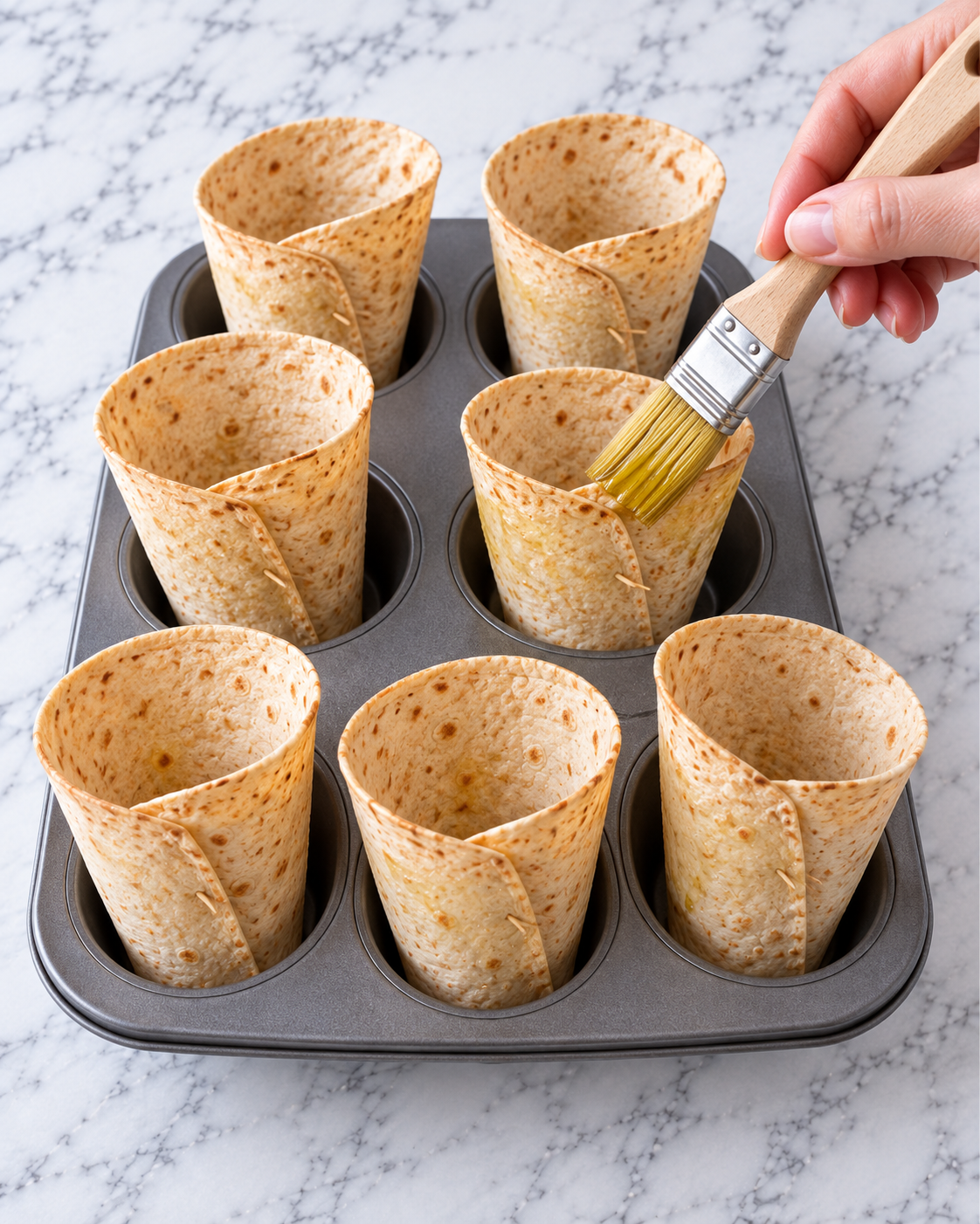Tortilla cones standing in a muffin pan while being brushed with olive oil
